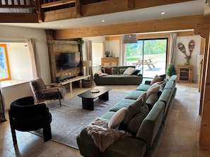 Ein geräumiges Wohnzimmer mit einer großen Eckcouch, einem flachbildfernseher an einer Holzwand, einem Holzesstisch und Blick ins Freie durch eine Schiebetür.