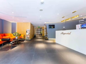 Ein Hotel-Empfangsbereich mit Empfangstresen, einer Sitzecke mit orangefarbenen Stühlen und einem an der Wand befestigten Fernseher.