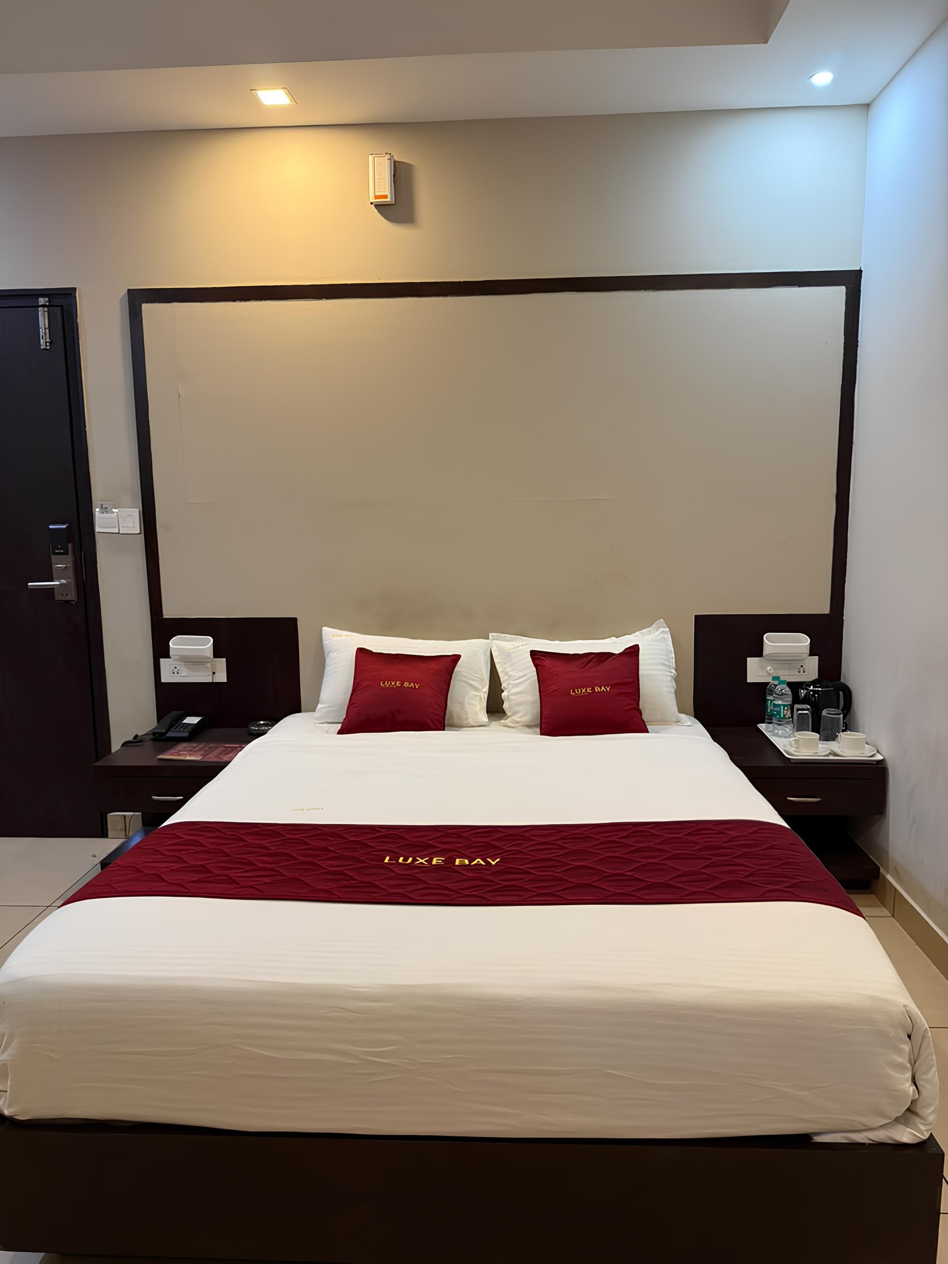 Habitación de hotel con una cama grande, dos mesitas de noche, un termostato en la pared y una puerta.
