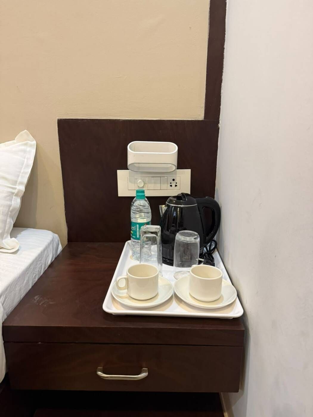 Una habitación de hotel con cama, una mesita de noche con bandeja que contiene dos tazas, una botella de agua, un vaso y una tetera.