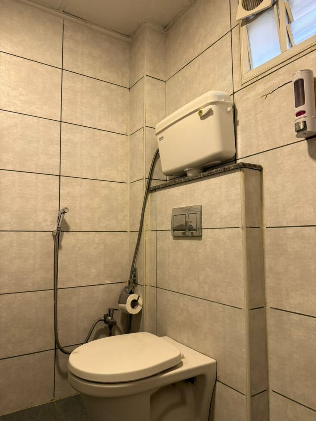 Un baño pequeño con azulejos, un inodoro, una manguera de ducha y un dispensador de papel higiénico.