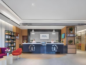 Un espace d’accueil d’hôtel au design moderne, comprenant un comptoir d’accueil, un grand écran affichant le nom de l’hôtel, et un coin salon avec un fauteuil rouge et un canapé violet.