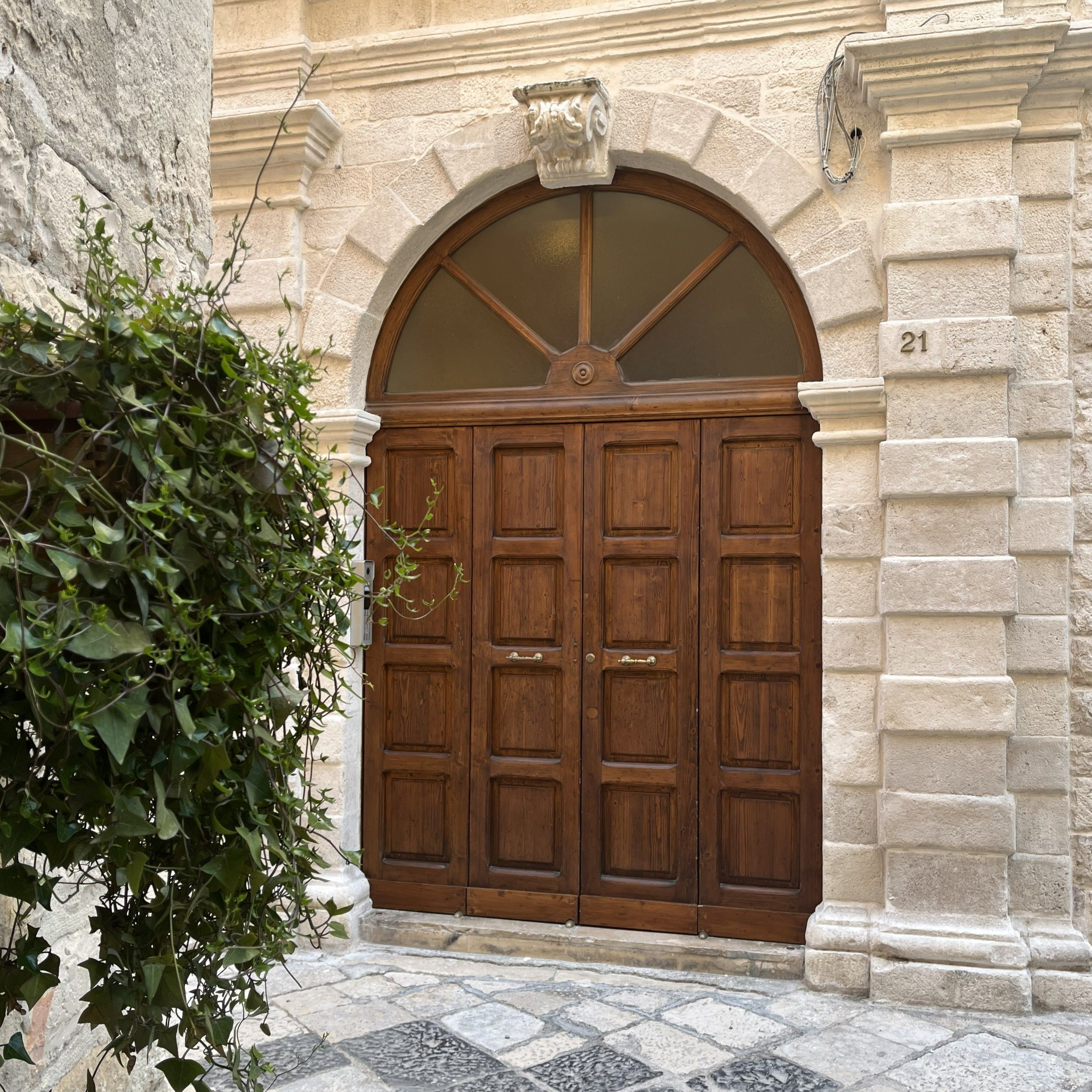 A large wooden double door with a semi-circular arch above it, set in a stone building with a number '21' visible on the right.