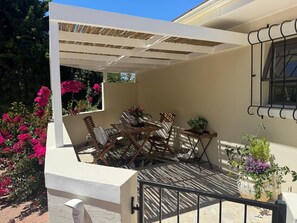 A covered outdoor seating area with a table and chairs, surrounded by potted plants and flowers.