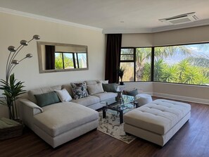 A living room with a sectional sofa, a glass coffee table, and a large window with a view of greenery.
