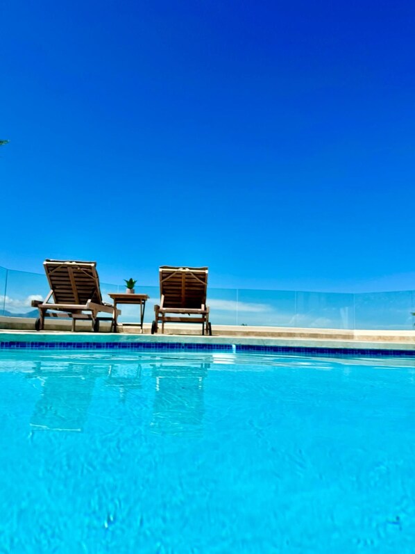 A clear blue pool with two wooden lounge chairs and a small table with a plant.