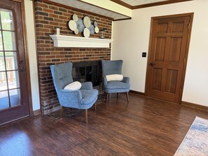 A room with a brick fireplace, two armchairs, a wooden door, and a wooden floor.