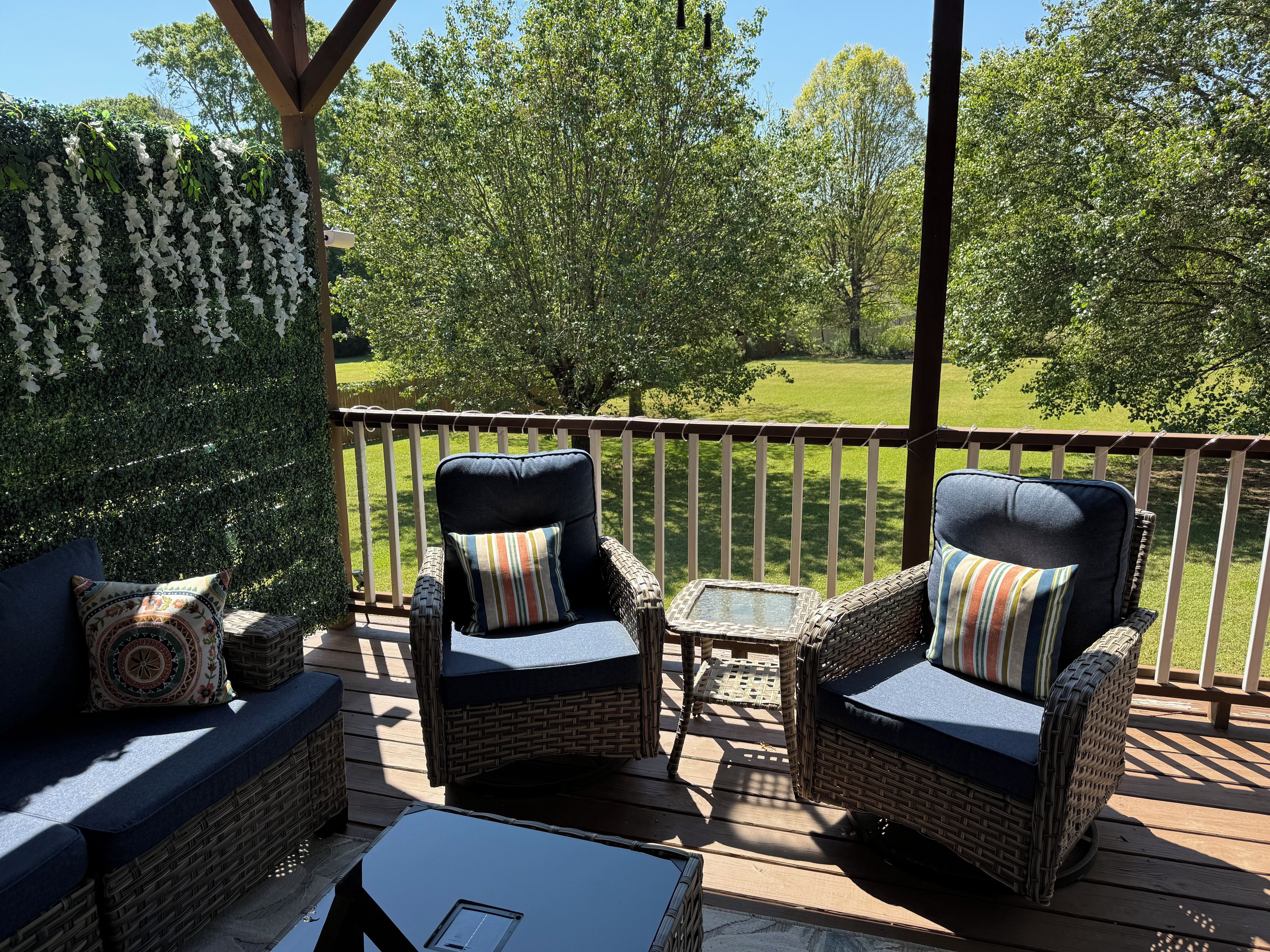 Eine Terrasse mit Korbmöbeln, darunter zwei Stühle und eine Couch, mit Blick auf einen Rasen und Bäume.