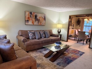 A living room with a brown sofa, patterned cushions, a glass coffee table, and a colorful rug.