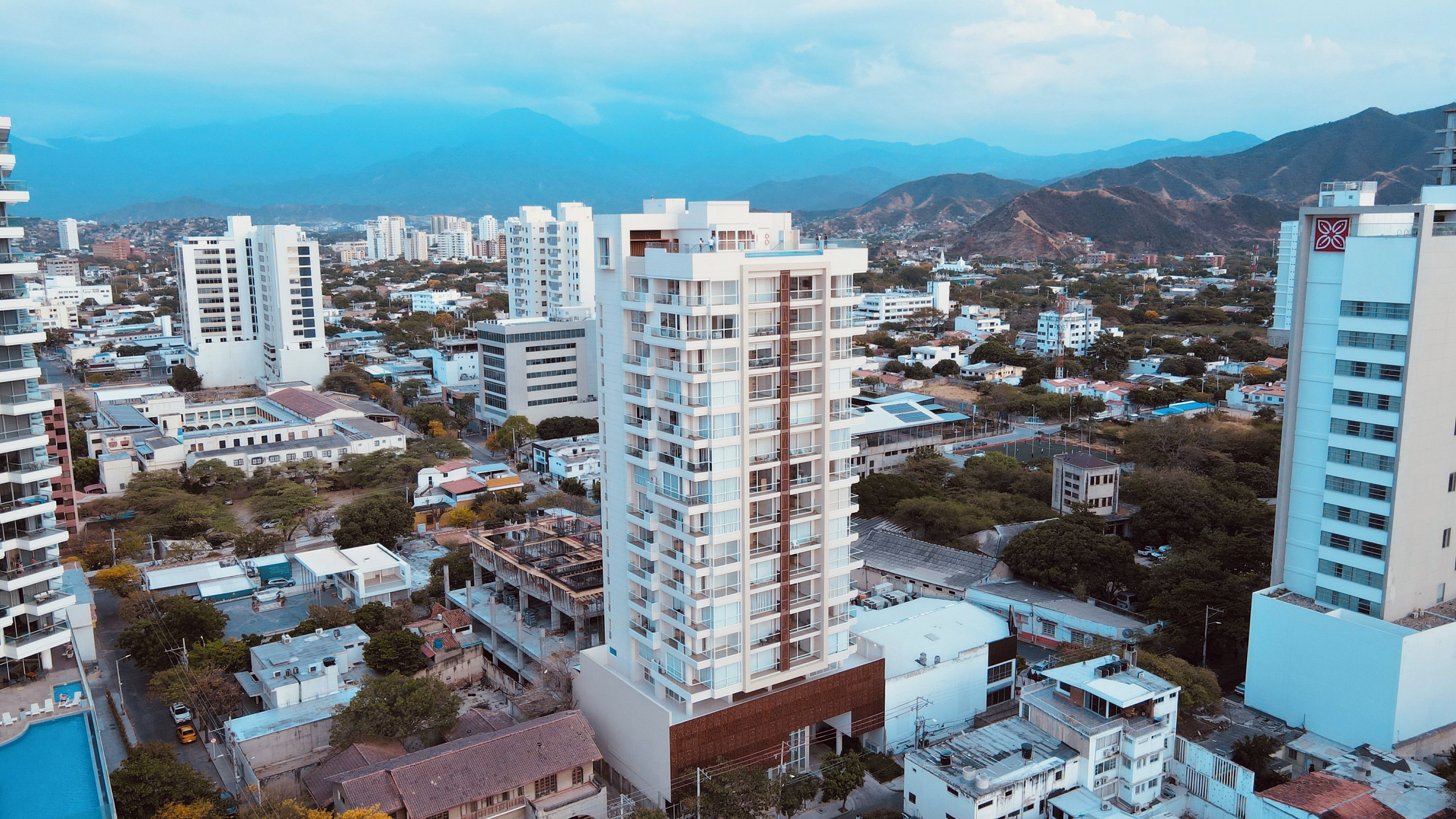 Un edificio de gran altura con varios pisos, rodeado de otros edificios y un paisaje montañoso al fondo.