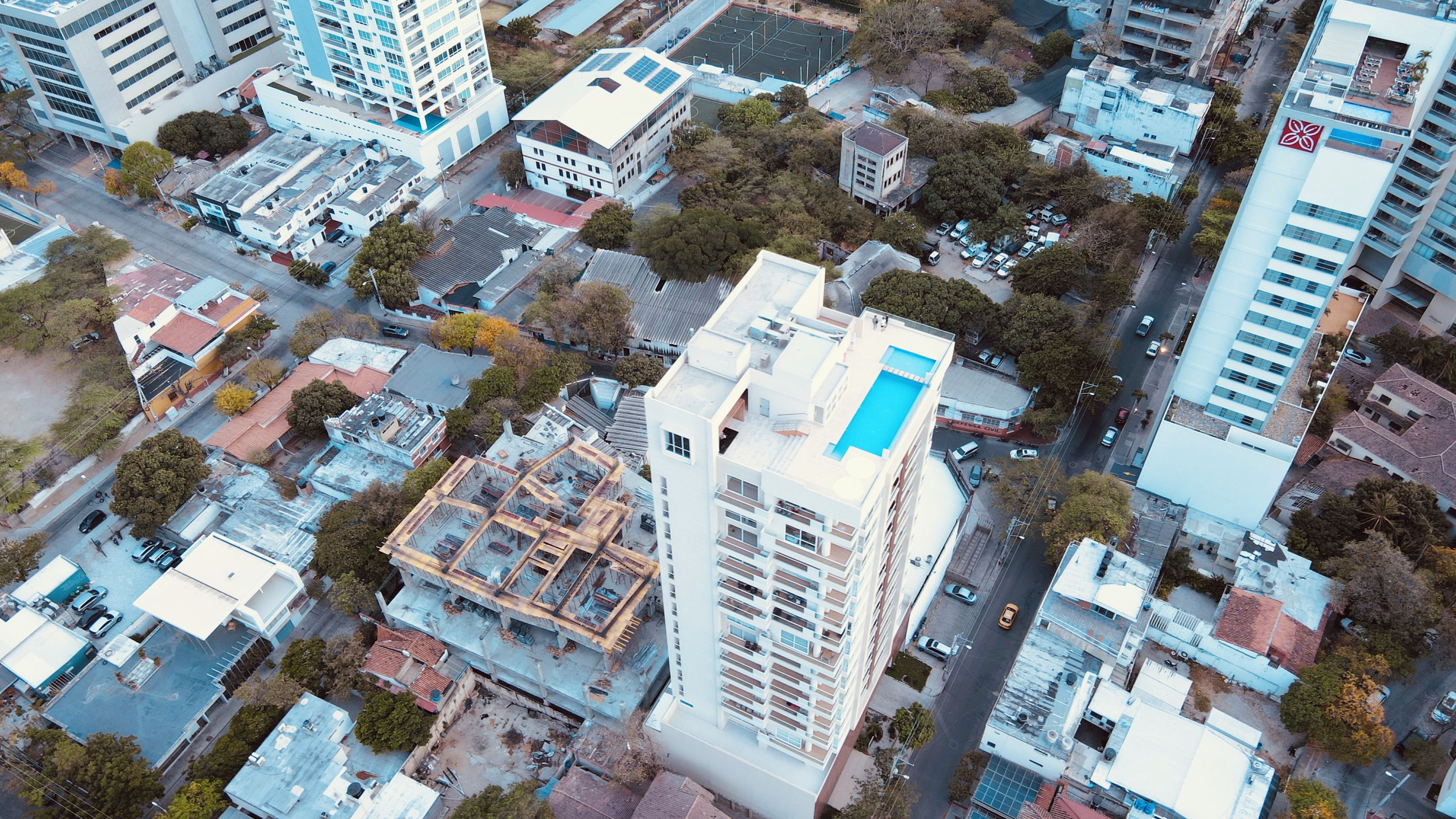 Vista aérea de un paisaje urbano con varios edificios, incluyendo un edificio blanco destacado con techo azul y un autobús rojo y blanco.