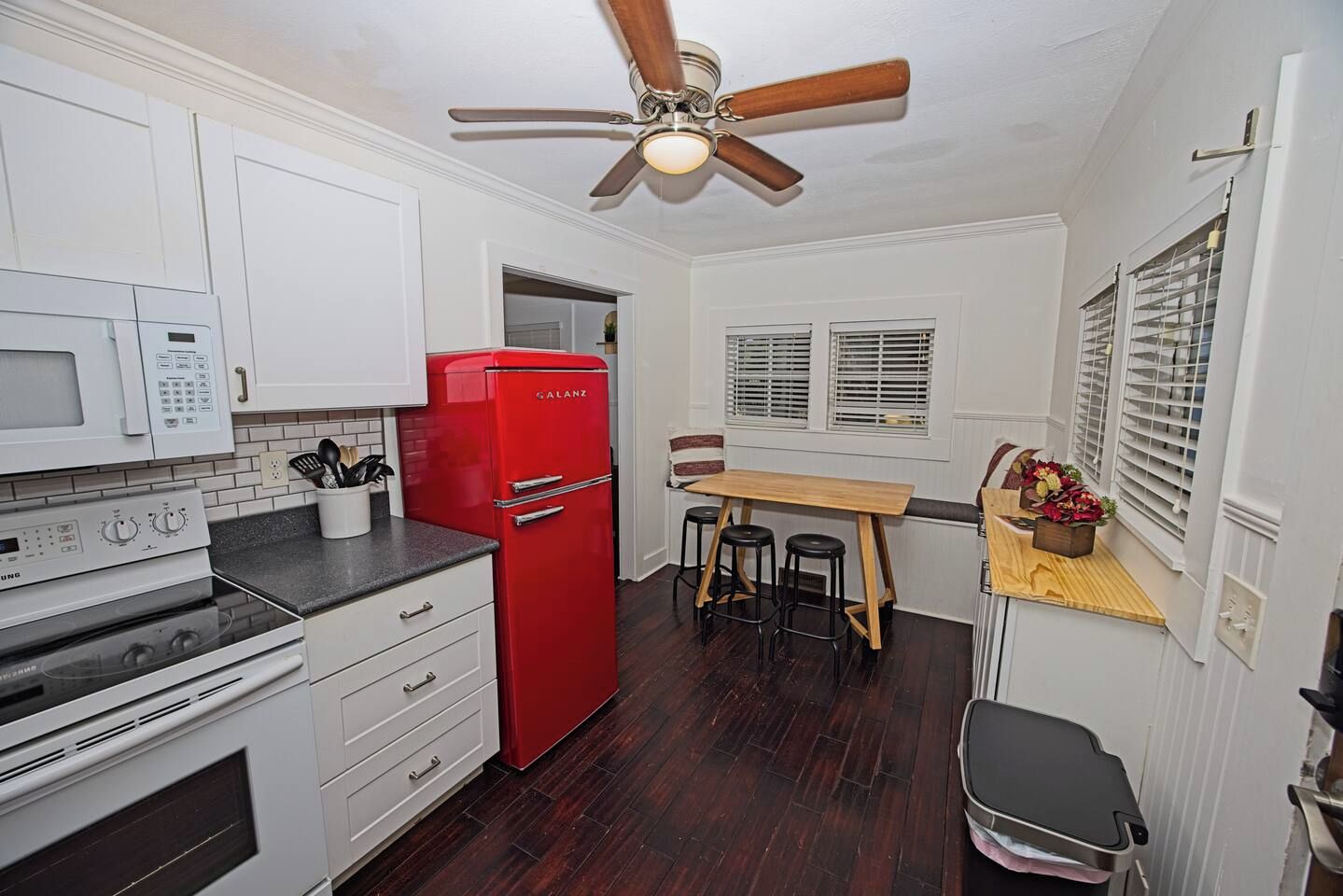 A compact kitchen with white cabinets, a red refrigerator, a microwave, an oven, a ceiling fan, a dining table with stools, and a window with blinds.