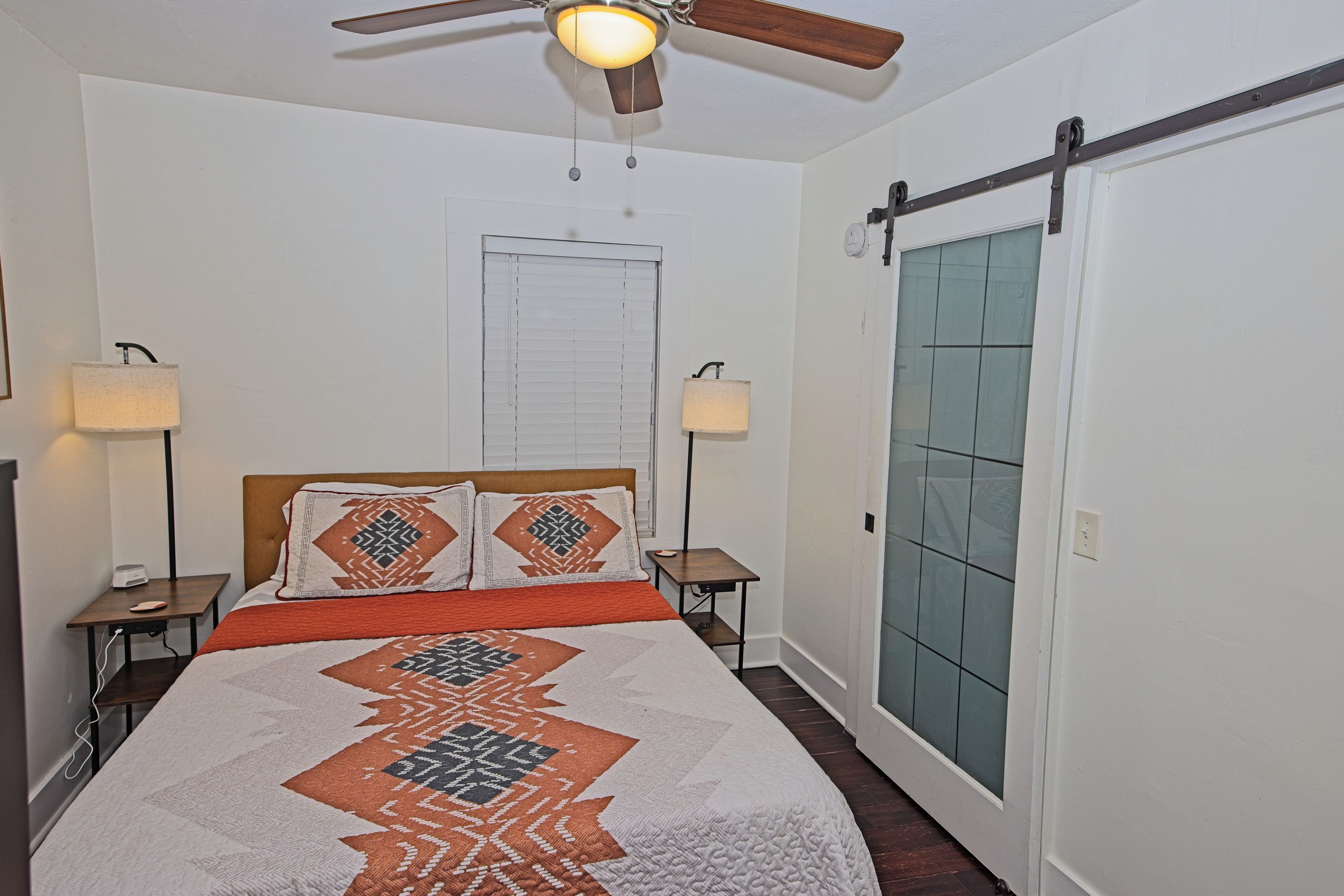 A bedroom with a bed, two bedside lamps, a ceiling fan, a window with blinds, and a door with a sliding mechanism.