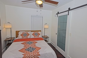 A bedroom with a bed, two bedside lamps, a ceiling fan, a window with blinds, and a door with a sliding mechanism.