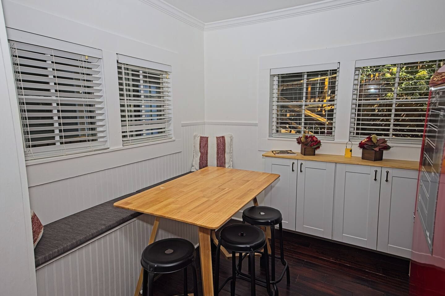 A compact kitchen with a wooden table and bar stools, white cabinetry, and a window with blinds.