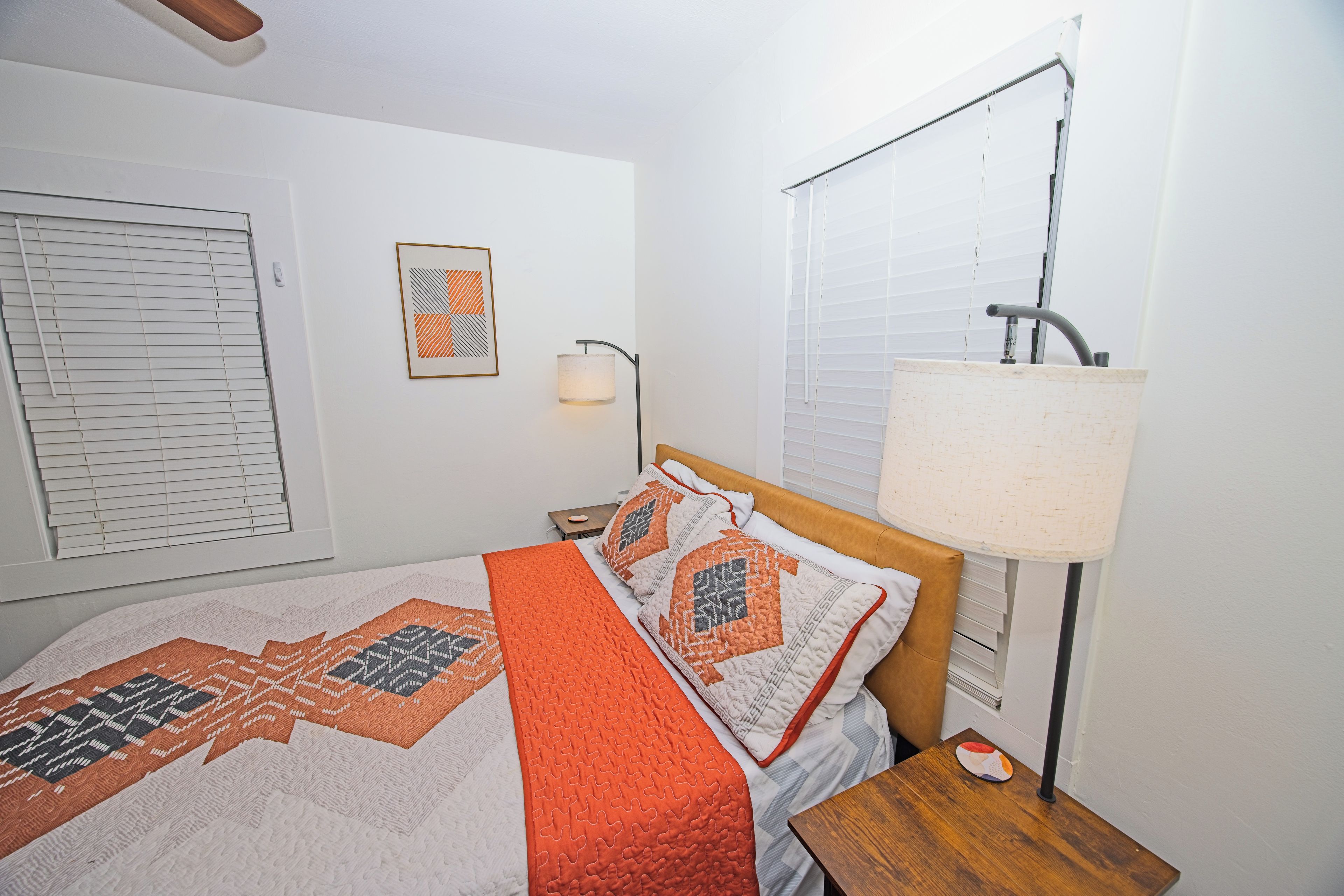 A bedroom with a bed, a bedside table, a lamp, a window with blinds, and a wall-mounted picture.