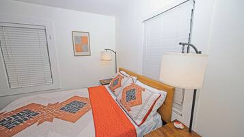 A bedroom with a bed, a bedside table, a lamp, a window with blinds, and a wall-mounted picture.