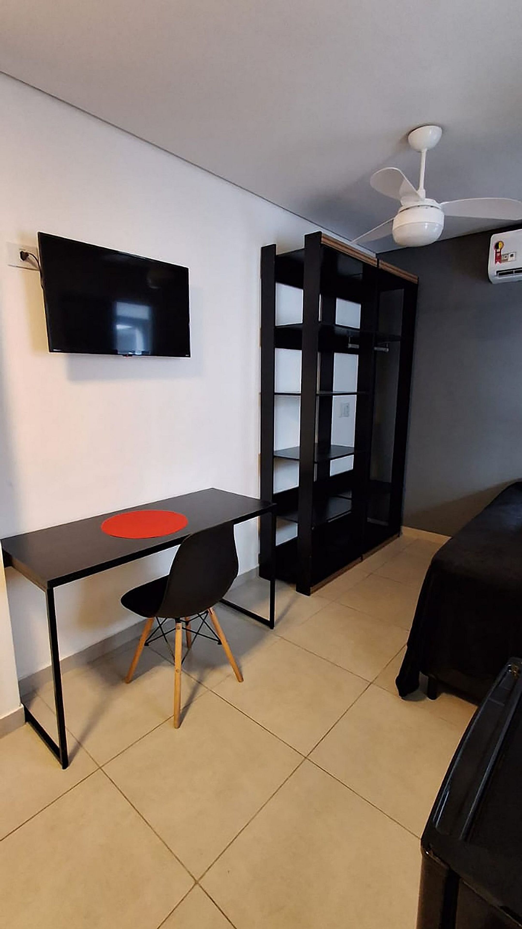 Et kompakt værelse med et fladskærms-tv på væggen, et sort skrivebord med en rund, rød genstand, en sort stol og en sort reol.