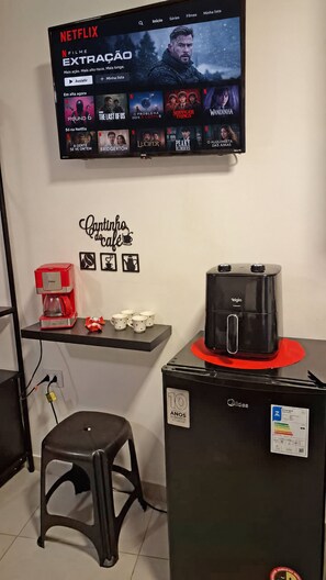 A room with a television displaying a Netflix interface, a coffee machine, a refrigerator, and a stool.