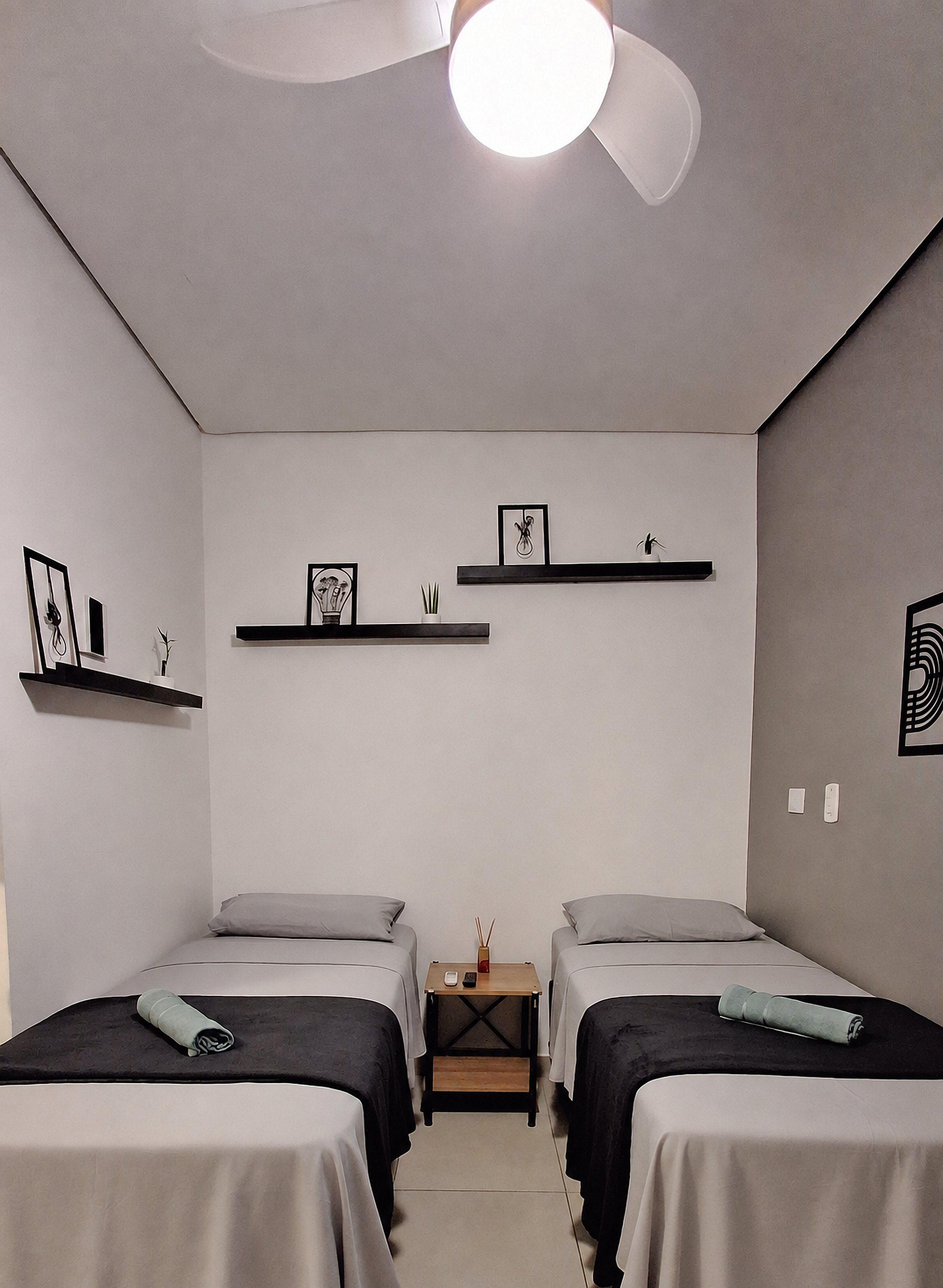 A room with two beds, a bedside table, a nightstand, and wall-mounted shelves with framed pictures.