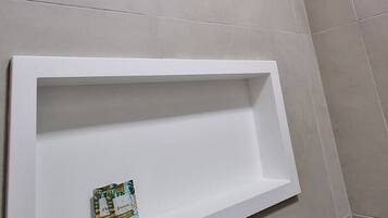 A bathroom with a shower, a white shelf, and tiled walls.