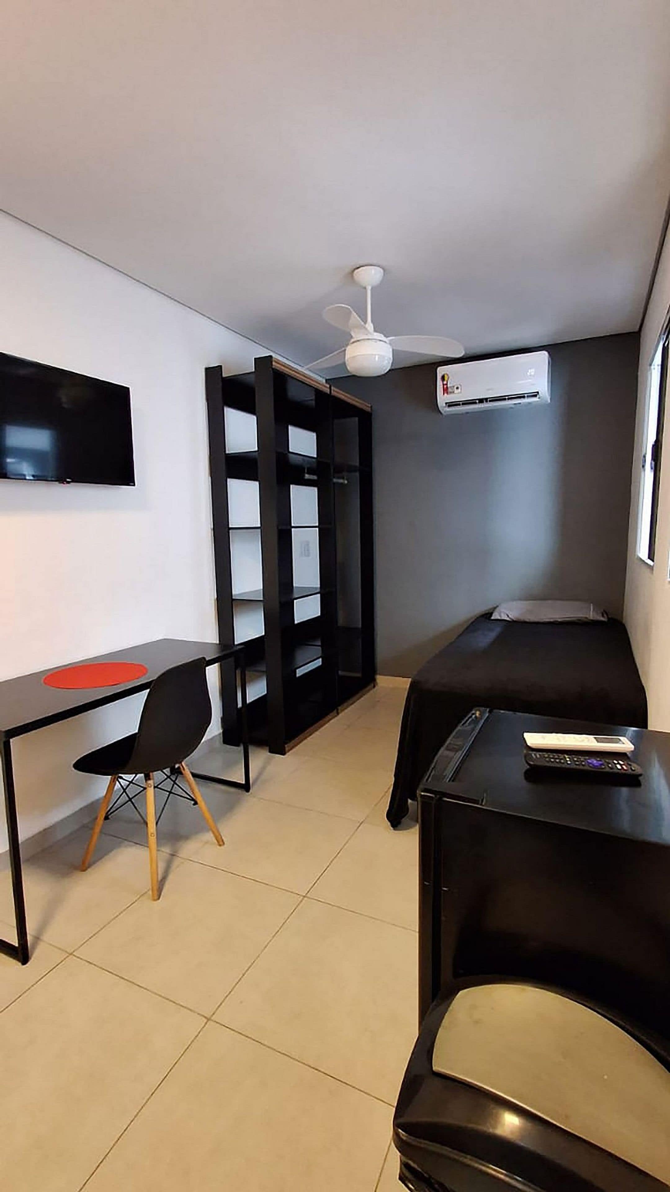 Quarto compacto com cama, escrivaninha, cadeira, televisão e ventoinha no teto.