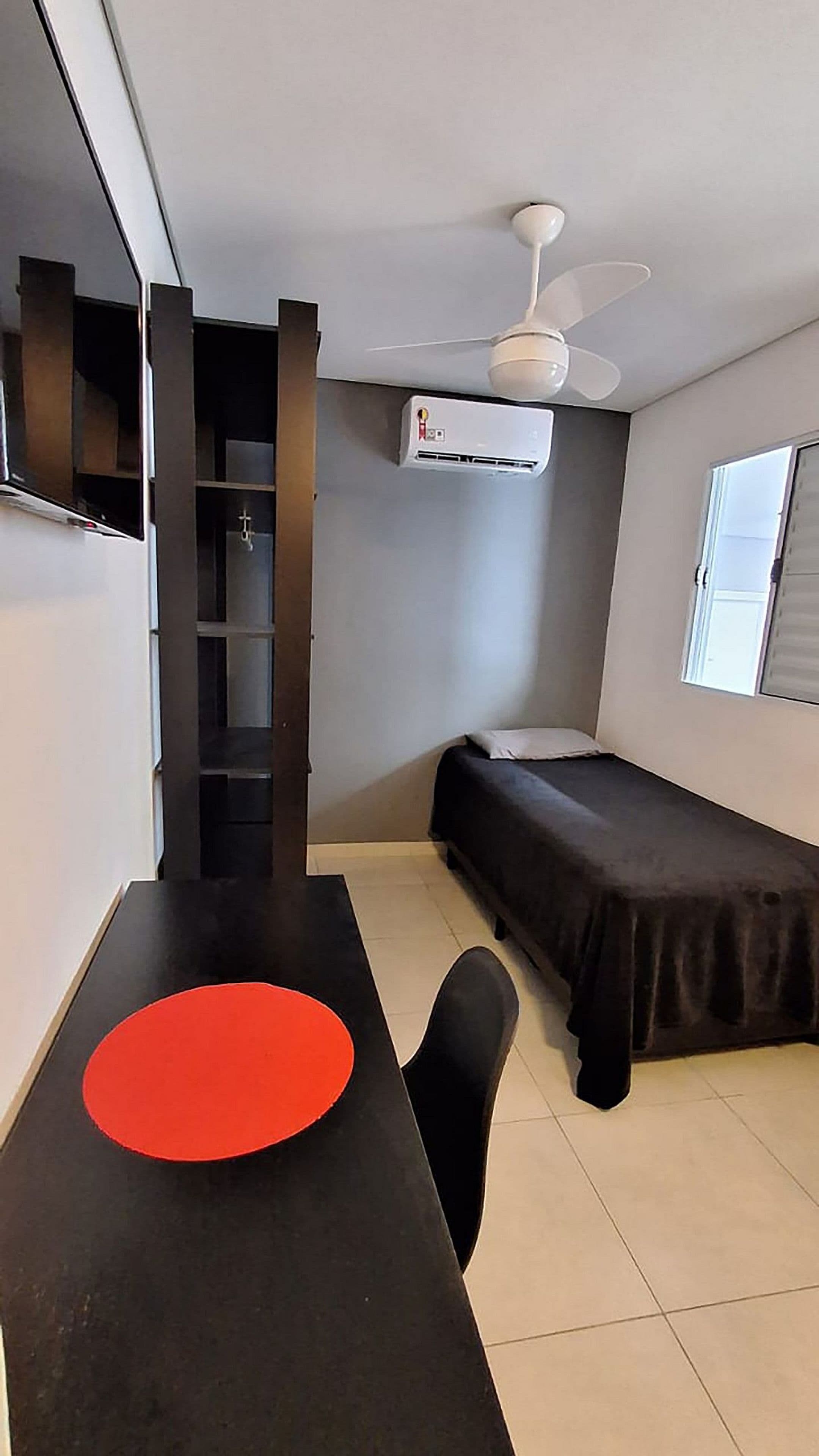 Een compacte kamer met een bed, een bureau met stoel, een plafondventilator en een airconditioning.