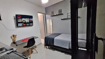 Une chambre moderne avec un lit, un bureau équipé d’un ordinateur, un téléviseur fixé au mur et une porte donnant sur une autre pièce.