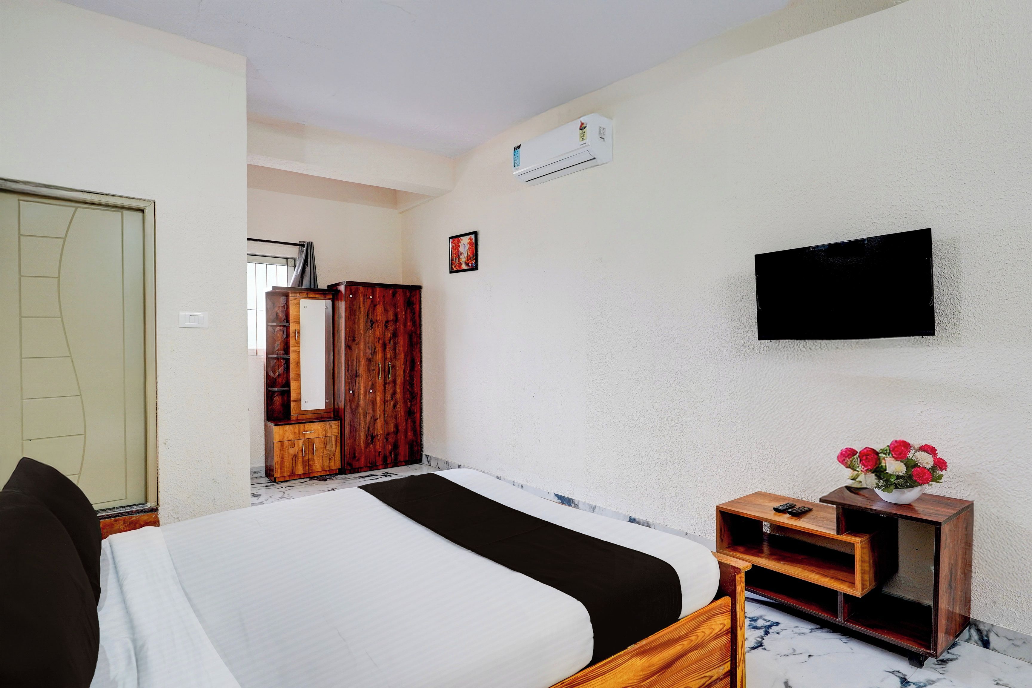 Habitación de hotel con cama, televisor montado en la pared, armario de madera y una mesita con un florero.