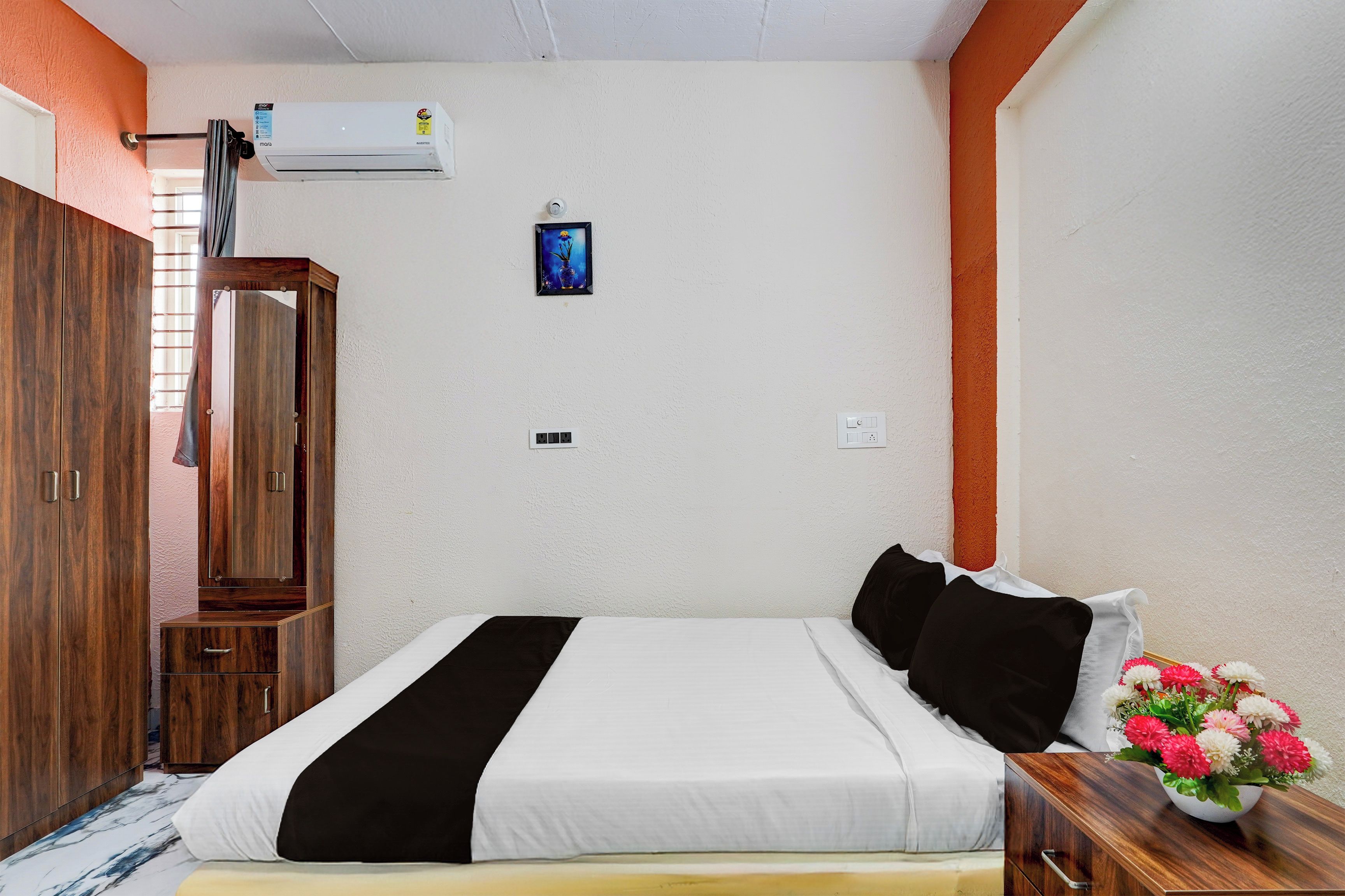 Una habitación de hotel con una cama, un armario de madera, un equipo de aire acondicionado, un interruptor de luz en la pared y un pequeño cuadro enmarcado.