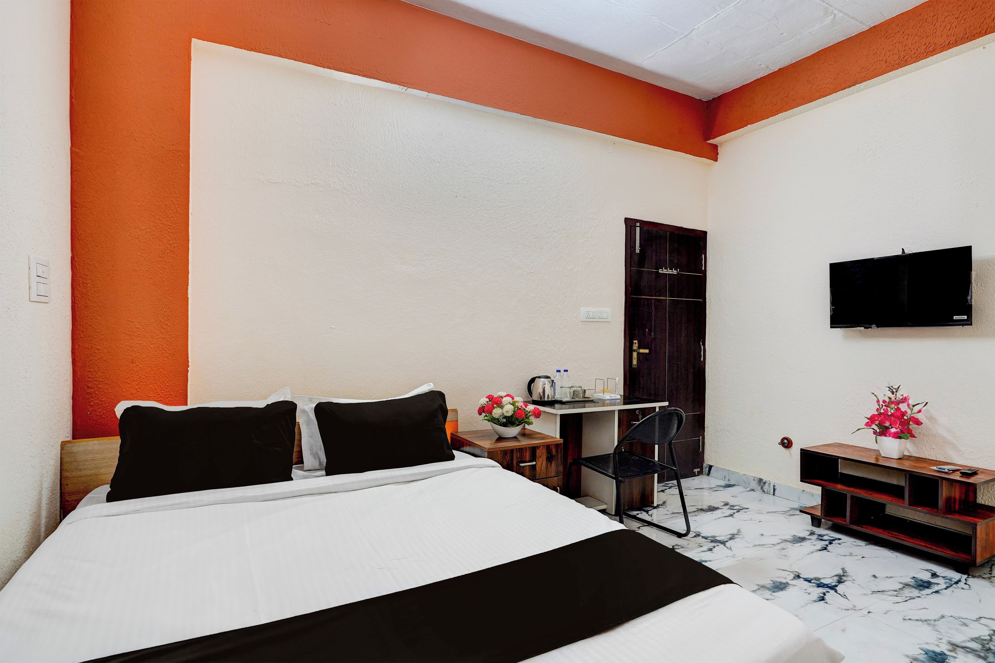 A hotel room with a bed, a desk, a chair, a TV, and a small table with flowers.