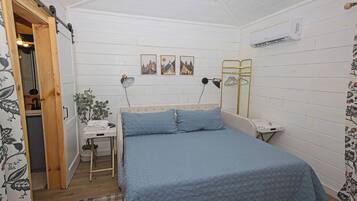 A bedroom with a bed, bedside table, lamp, wall art, and an air conditioning unit.