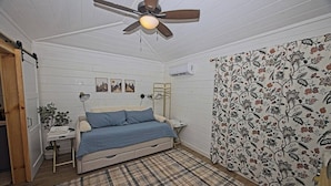 Een slaapkamer met een bed, een plafondventilator, een nachtkastje, een lamp, een wandairconditioner en een kamerbreed gordijn.