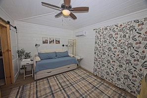 A bedroom with a bed, a ceiling fan, a nightstand, a lamp, a wall-mounted air conditioner, and a patterned curtain. - Mini Messer - Downtown, Walk to Shops (Blue Ridge)