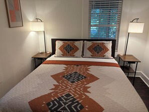 Een netjes opgemaakt bed met beddengoed met een patroon, twee bedlampen en een raam met jaloezieën.