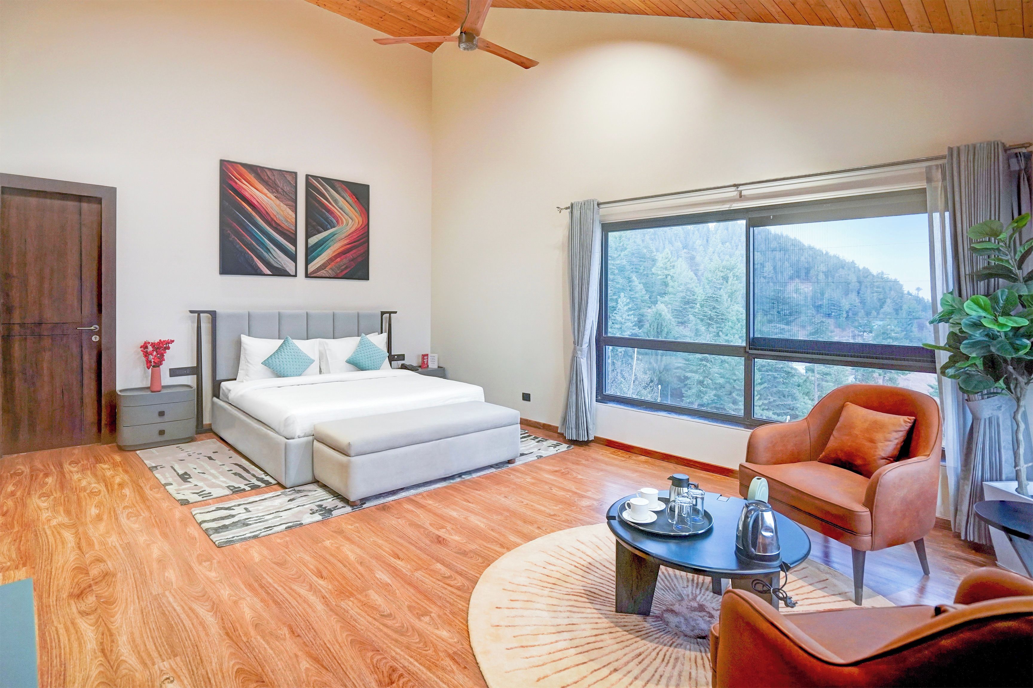 A bedroom with a bed, a coffee table, a chair, a window with a view of trees, and artwork on the wall.