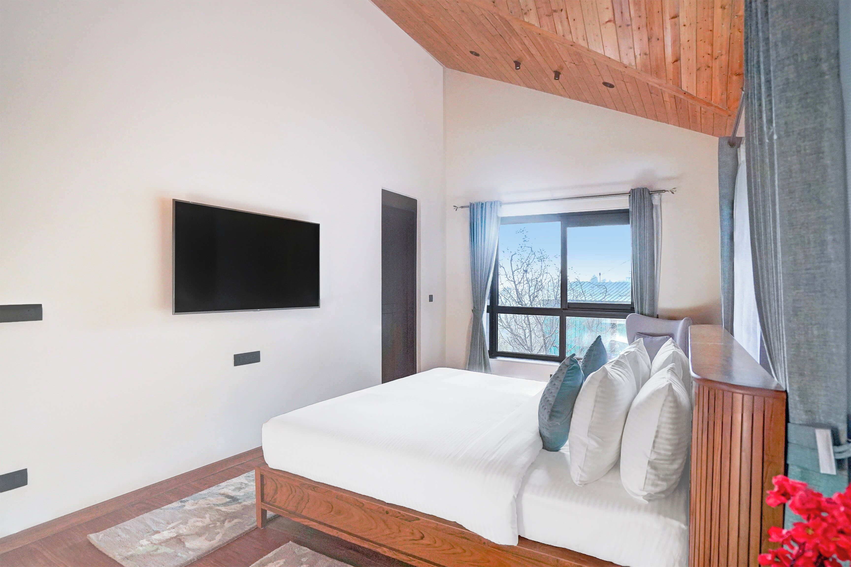 A bedroom with a bed, a flat-screen TV mounted on the wall, a window with curtains, and a view of trees outside.