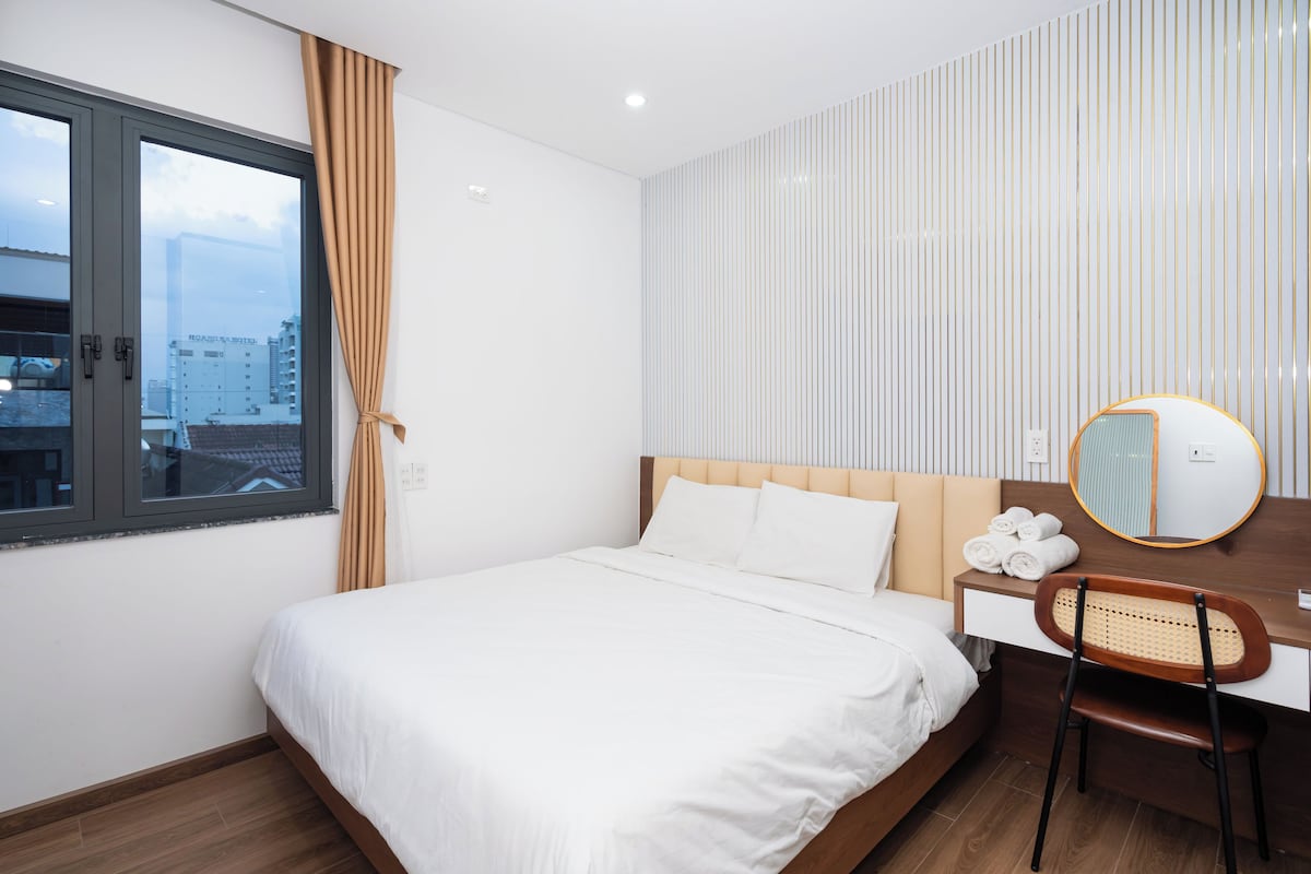 A hotel room with a bed, a desk with a chair, a mirror, and a window with curtains.