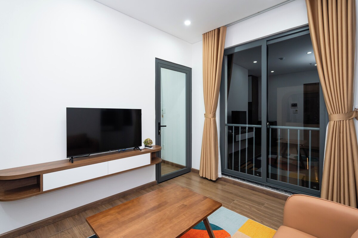A modern living room with a flat-screen TV mounted on a wooden console, a sliding glass door leading to a balcony, and a colorful rug on the floor.
