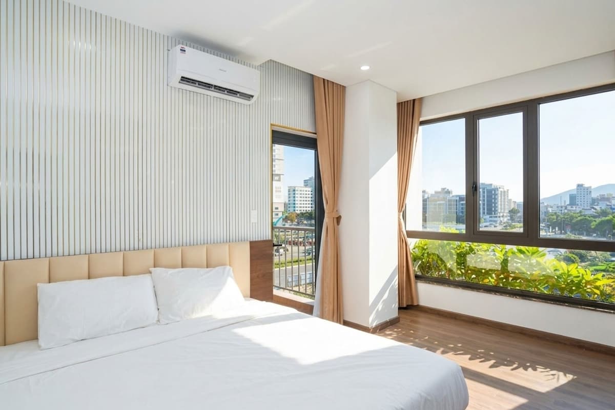 A hotel room with a bed, a window with curtains, a view of the city, and an air conditioning unit.