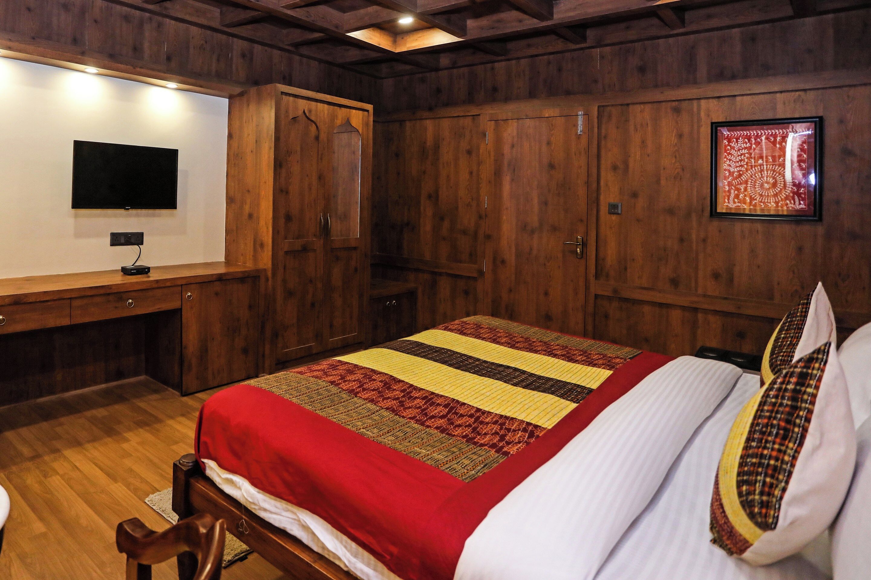 Un dormitorio con piso de madera, una cama con colcha a rayas roja y amarilla, una almohada blanca, una mesita de noche de madera, un televisor de pantalla plana montado en la pared y un cuadro enmarcado en la pared.