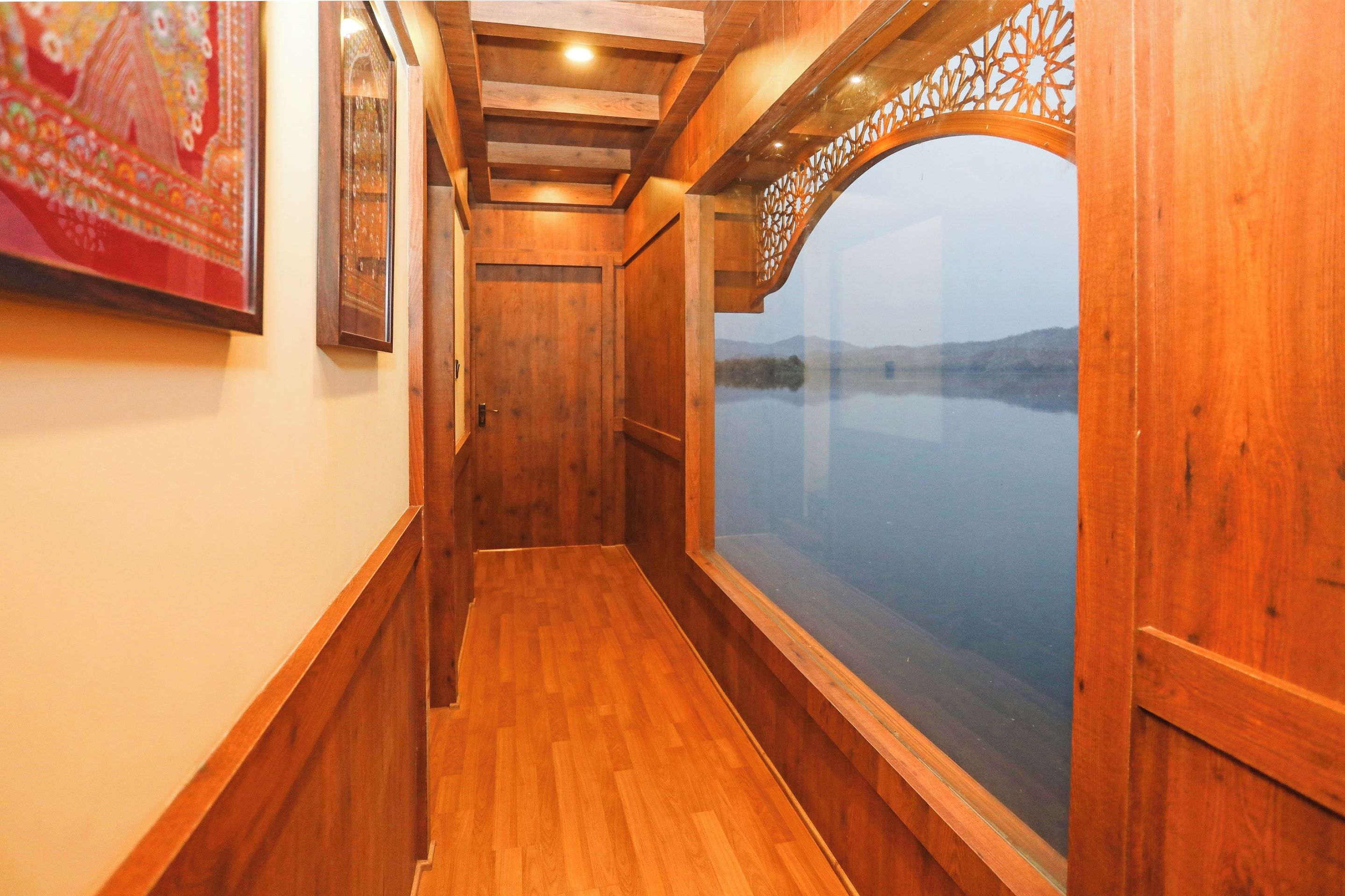 Un estrecho pasillo de madera con una puerta al final, una ventana con vista a una masa de agua y un cuadro en la pared.