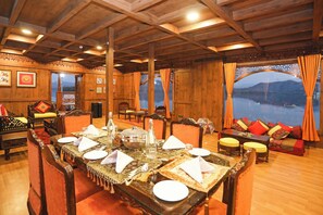 A dining area with a long table set for a meal, wooden walls, and a view of a body of water through large windows. - Ekta Houseboat Statue Of Unity Kevadia (Vadodara)