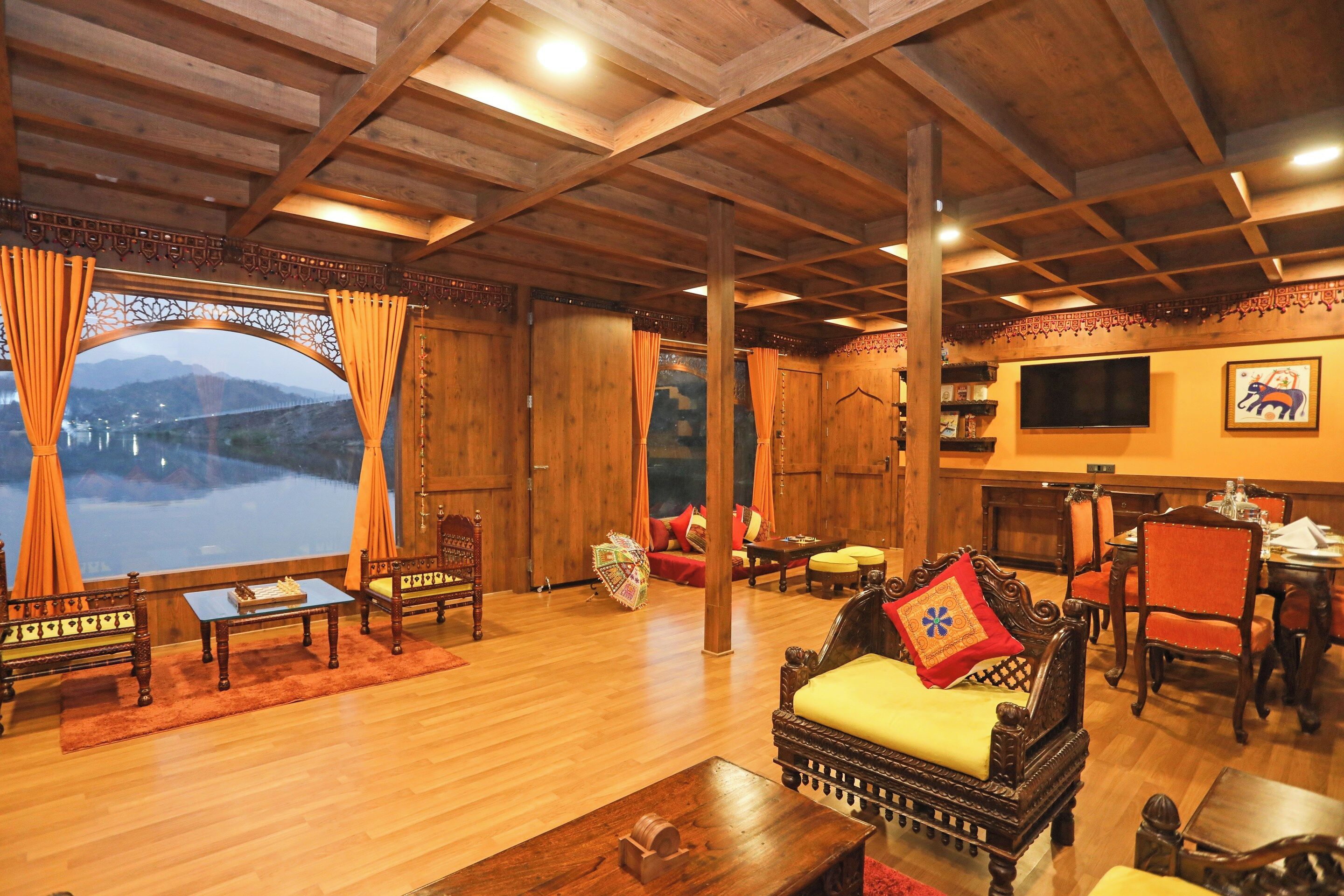 A spacious living area with wooden furniture, a large window with curtains, and a television on the wall.