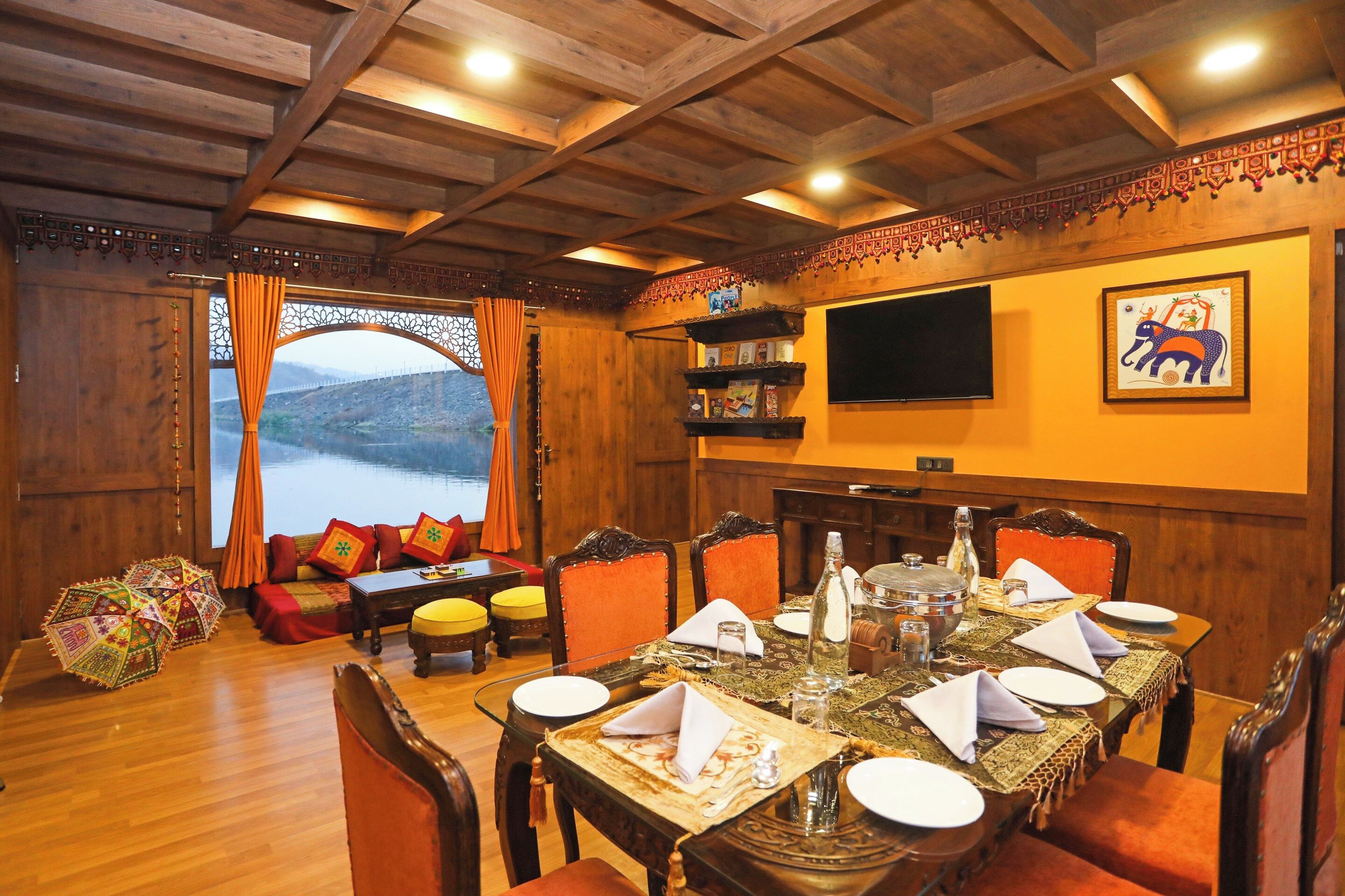 A dining area with a wooden ceiling, a large window with curtains, a television mounted on the wall, and a table set for dining.