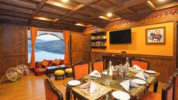 A dining area with a wooden ceiling, a large window with curtains, a television mounted on the wall, and a table set for dining.