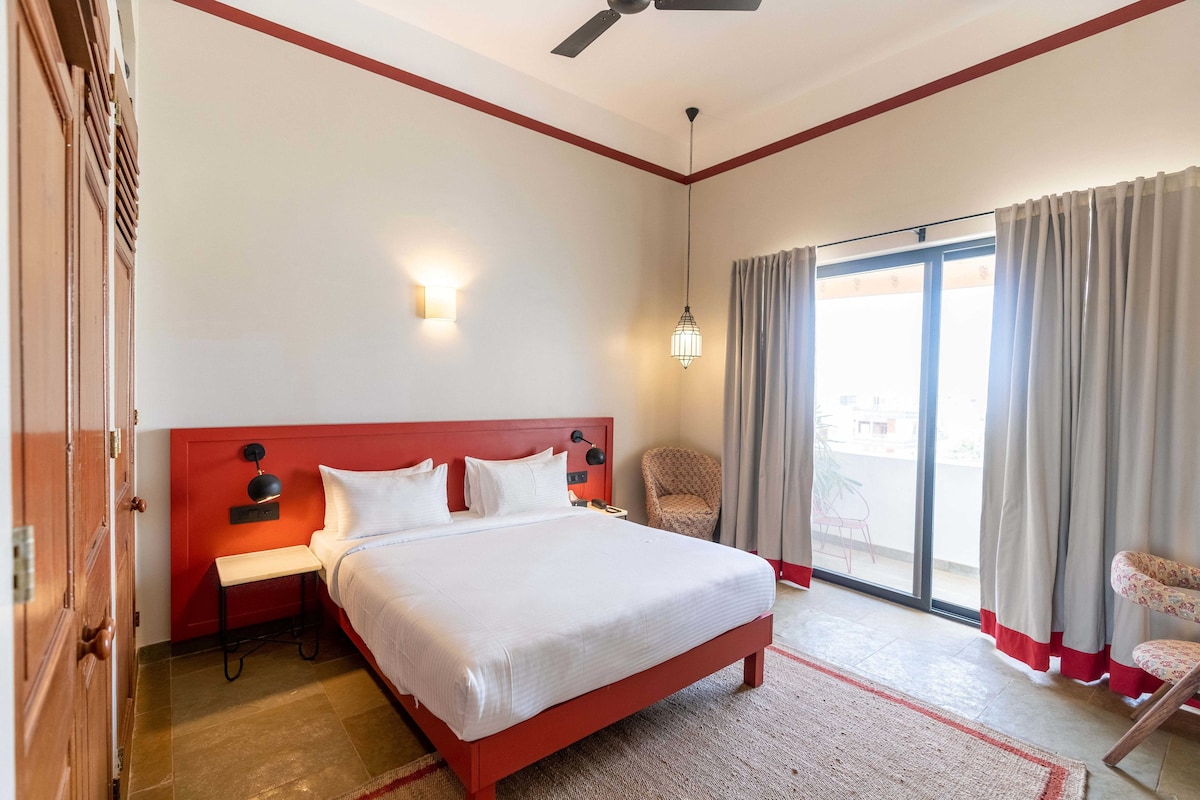 Ein Hotelzimmer mit einem Bett, einem Nachttisch, einem Sessel, einem Fenster mit Vorhängen und einem Deckenventilator.