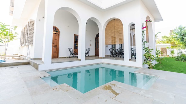 Ein Poolbereich mit klarem Pool, umgeben von weißen Wänden und Bögen, inklusive Außenmöbeln und Blick auf ein Wohngebäude.