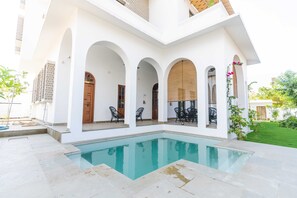 A pool area with a clear pool, surrounded by white walls and arches, with outdoor seating and a view of a residential building. - House of Jasol by DanCenter, Jaipur (Jaipur)