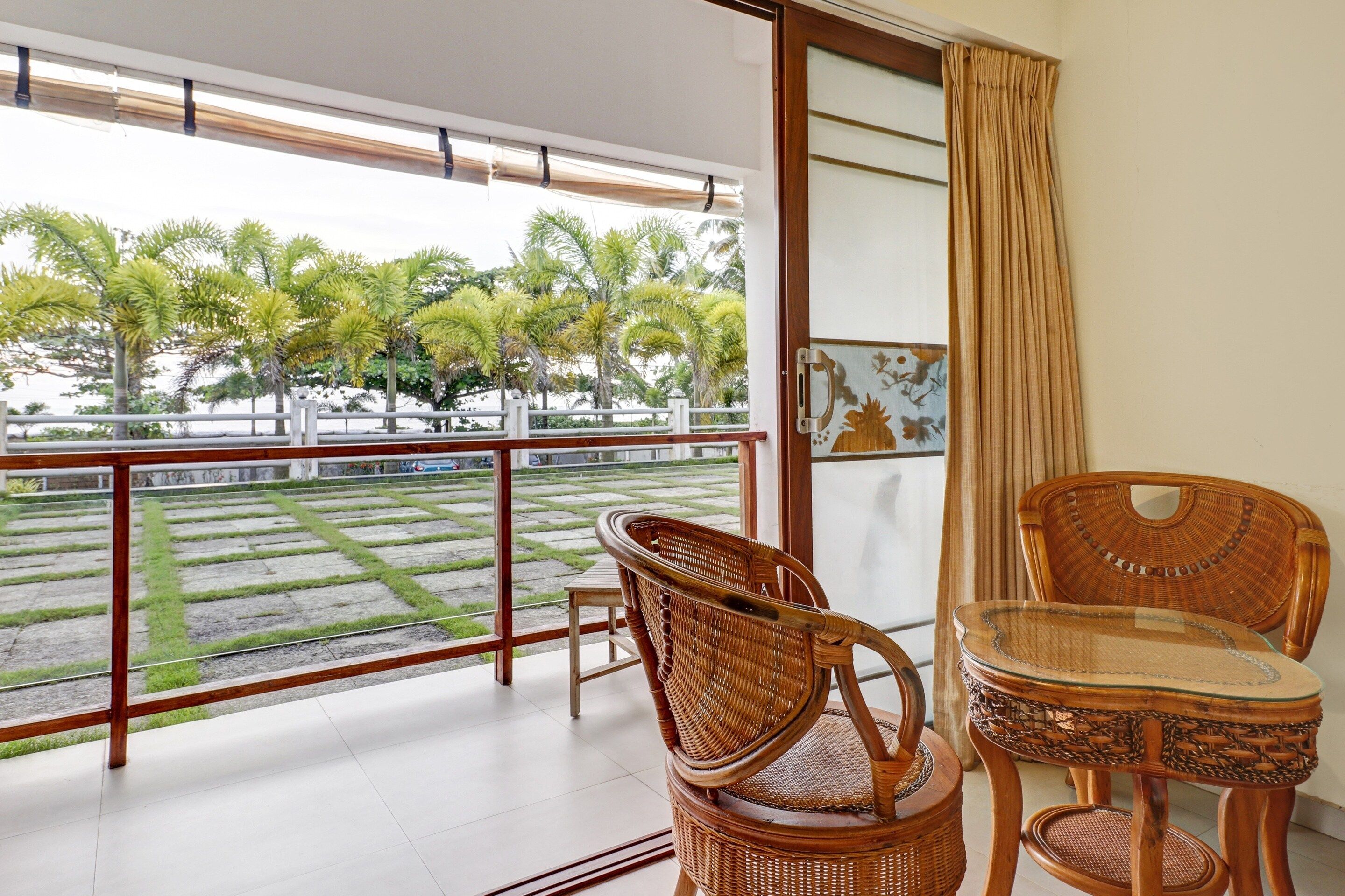 A balcony with wicker furniture, a glass table, and a view of a landscaped area with palm trees.
